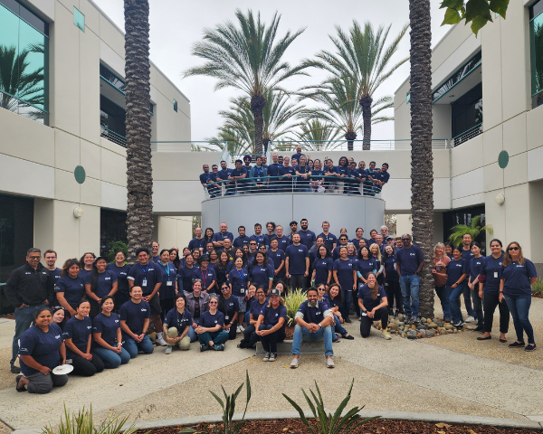 Un groupe de personnes portant des t-shirts de BD, des charlottes, des gants et des badges d'identification sont rassemblés dans un environnement de production ou de laboratoire intérieur, avec du matériel visible en arrière-plan.