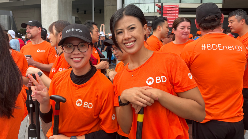 Duas mulheres vestindo camisas BD laranja vibrante, sorrindo em um evento de corrida de barcos.