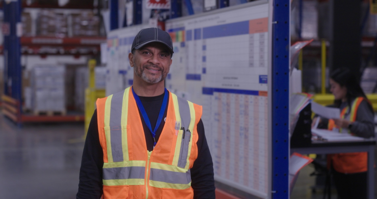 Indivíduo usando um colete de segurança laranja de alta visibilidade e um boné, em pé dentro de um armazém com prateleiras e gráficos ao fundo.