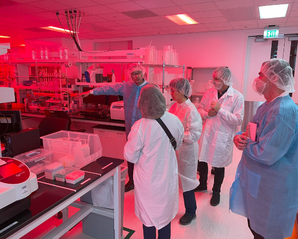 Un groupe de personnes en blouse blanche, charlotte et équipement de protection se tiennent dans un laboratoire éclairé par une lumière rouge. L'une d'elles pointe du doigt du matériel sur une paillasse, tandis que les autres observent.