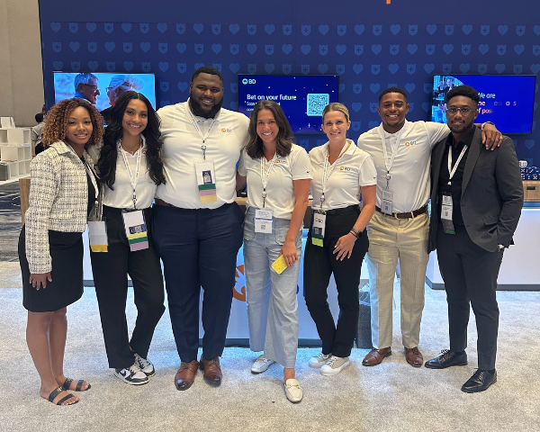 Un groupe de sept pose devant un grand écran numérique dans une salle de conférence ou d'événement. Elles sont vêtues de tenues professionnelles ou décontractées chics, certaines portant des chemises blanches assorties avec des insignes et des cordons.