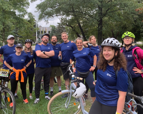 Um grupo de indivíduos reuniu-se ao ar livre, ao lado de bicicletas, todos vestindo camisas BD idênticas, com árvores e edifícios ao fundo.