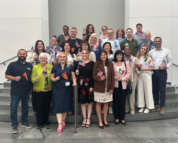 Um grande grupo de indivíduos reunidos em uma escadaria interna, cada um segurando uma pequena bandeira de mão de diversos países.
