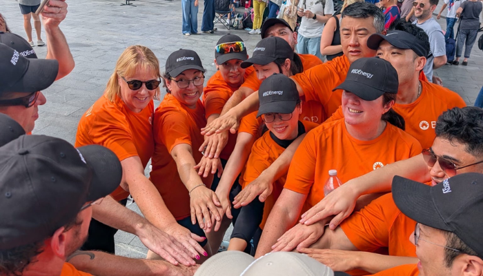 BD associates in orange event shirts stand in a team huddle with hands stacked together during an outdoor group activity.