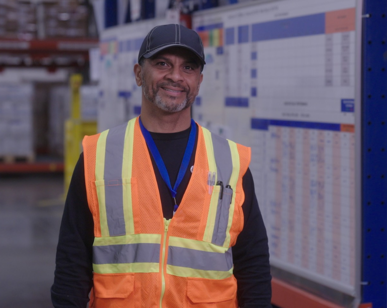 Individual wearing a high‑visibility orange safety vest and cap standing inside a warehouse with shelving and charts in the background.