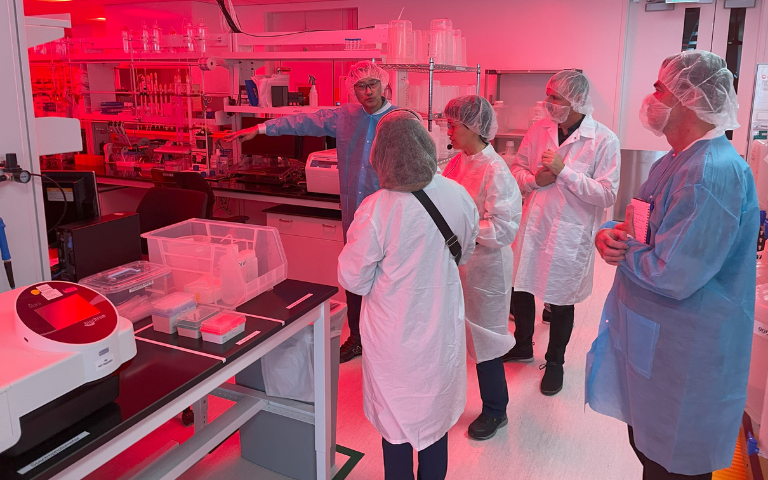 Grupo de indivíduos vestindo jalecos e trajes de proteção durante uma visita guiada no interior de um laboratório iluminado por luz vermelha, com uma pessoa apontando para um equipamento sobre uma bancada.