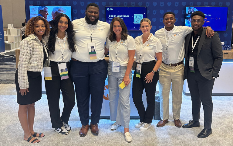 Um grande grupo de indivíduos reunidos em um estande de convenção ou exposição, com a marca BD exibida em telas ao fundo.