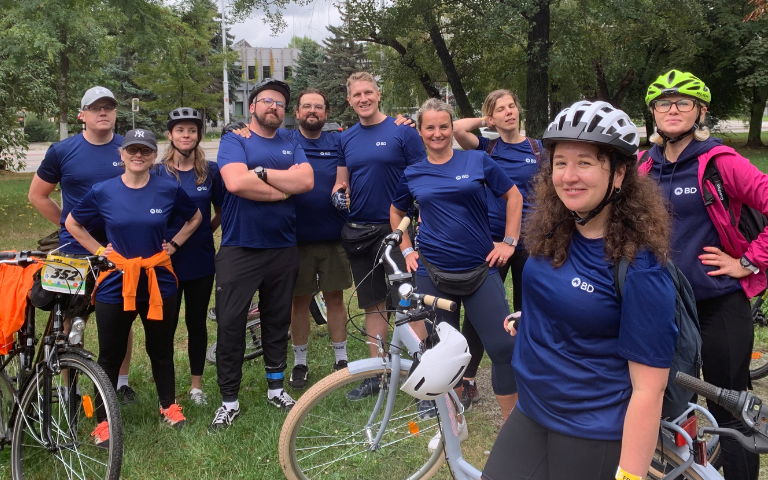 A group of individuals gathered outdoors beside bicycles, all wearing matching BD shirts, with trees and buildings in the background.