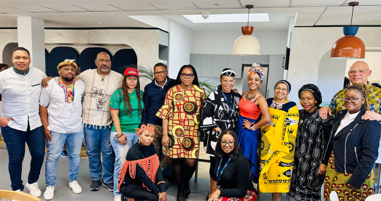 Group of adults standing together indoors by large windows, wearing colorful patterned clothing.