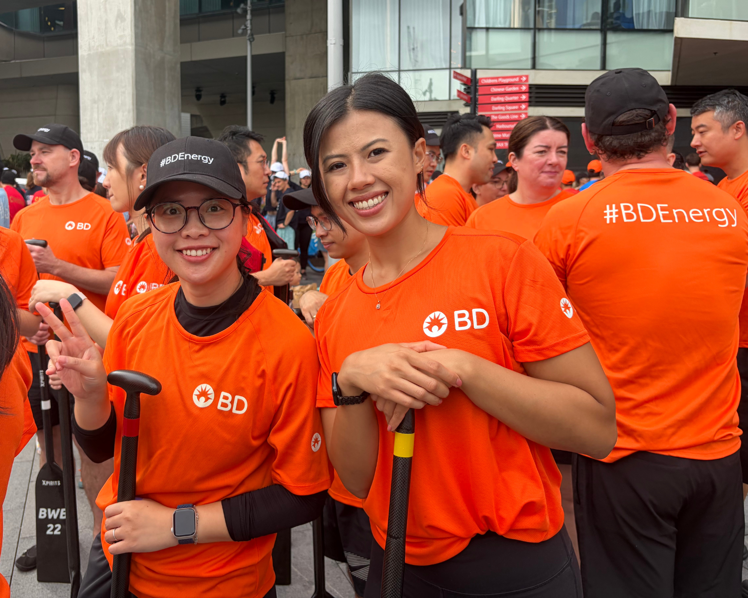 Two women wearing bright orange BD shirts smiling at a boat race event