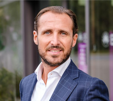 A portrait-style photo of an individual smiling into the camera, outside, wearing a patterned navy blazer and white collared top.