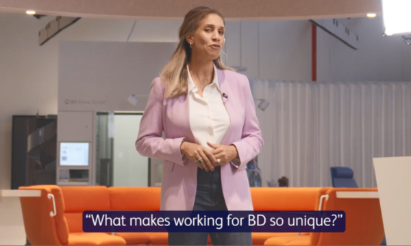 A lady with light hair, wearing a light purple blazer and white blouse, smiling enthusiastically at the camera.