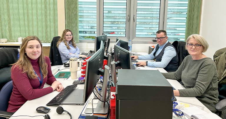 Four individuals seated at desks in a bright office with light-colored flooring, green vertical blinds, and large windows. Each workstation has multiple computer monitors, office supplies, and personal items arranged on the desks.