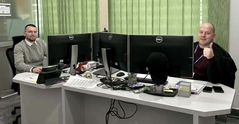 Two individuals seated at separate desks with multiple computer monitors in an office featuring green vertical blinds and a glass wall. Various office supplies, documents, and personal items are arranged on the desks