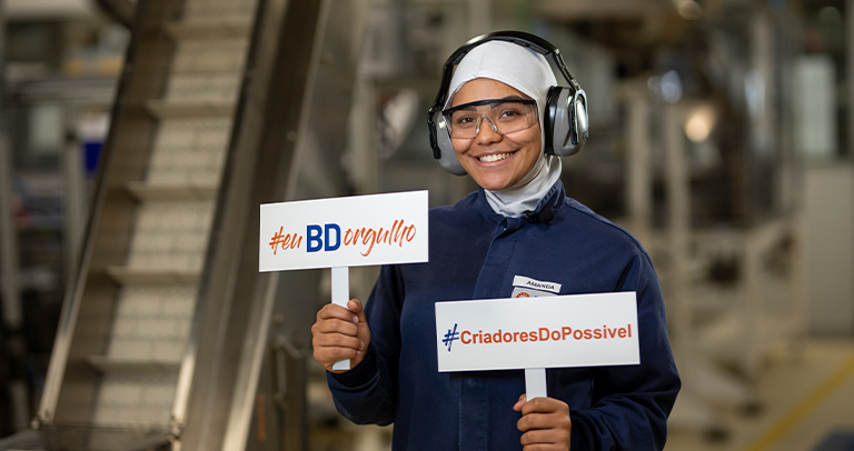 Individual in protective industrial attire holding two signs that read ‘#euBDorgulho’ and ‘#CriadoresDoPossivel,’ standing in a manufacturing environment with machinery and equipment in the background. The individual is wearing safety earmuffs, clear prot