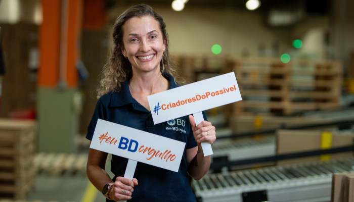 BD associate holding branded signs inside a manufacturing facility near conveyor equipment.
