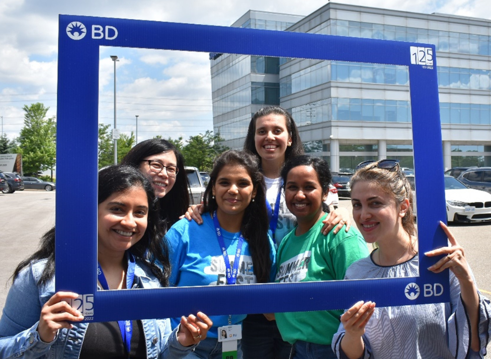 Un groupe de six personnes posant à l’extérieur devant un immeuble de bureaux vitré de plusieurs étages, tenant un grand cadre photo bleu à l’image de BD. Le cadre affiche le logo BD et un emblème « 125 ans » dans les coins supérieurs.