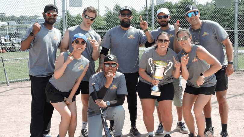 Un groupe de neuf personnes posant ensemble sur un terrain de baseball, avec une clôture à mailles et des arbres en arrière‑plan.