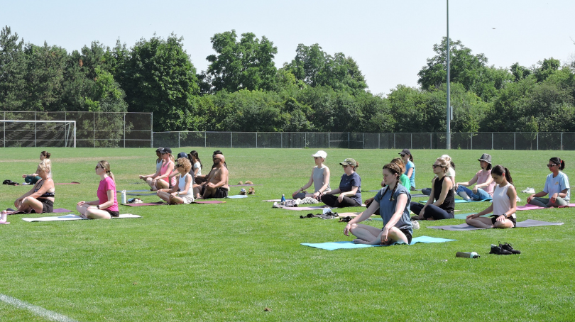 Un groupe de personnes assises sur des tapis de yoga disposés en rangées sur un terrain extérieur engazonné. Chaque personne est assise face vers l’avant, avec des arbres, une clôture et un ciel dégagé visibles en arrière‑plan.