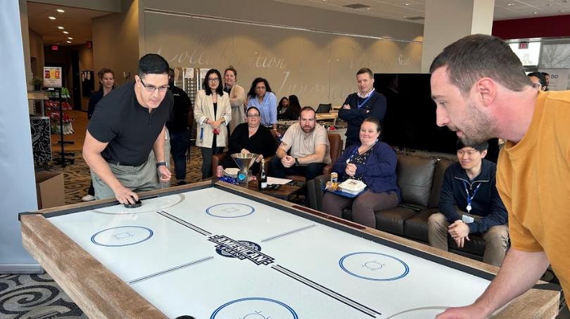 Deux personnes jouant à une partie de hockey sur coussin d’air sur une table grandeur nature dans un espace commun de bureau, tandis qu’un groupe de collègues est assis et debout autour pour regarder.