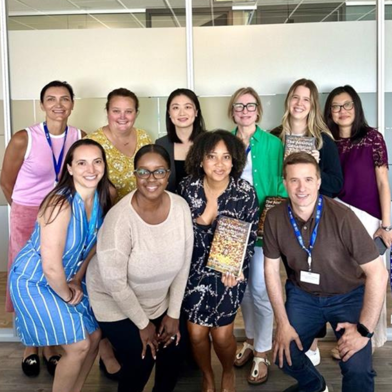 Un groupe de onze personnes posant ensemble dans un espace de bureau, avec des cloisons vitrées en arrière‑plan. Plusieurs personnes tiennent des livres, et le groupe est disposé en deux rangées, certaines assises et d’autres debout.