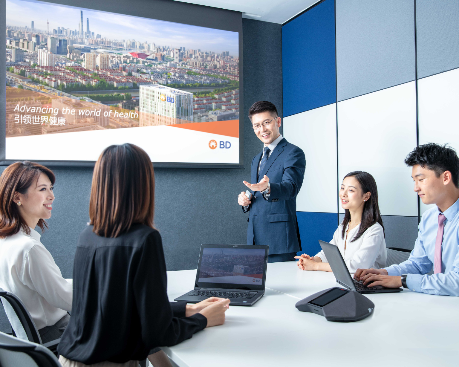 Five individuals seated and standing around a conference table in a modern meeting room, using laptops and looking toward a wall‑mounted presentation screen. The screen displays an image of a cityscape with a BD building.