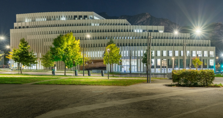 Nighttime exterior view of a large modern building with tiered rectangular architecture, illuminated by streetlights and surrounded by trees and landscaped paths, with mountains faintly visible in the background under a dark sky.