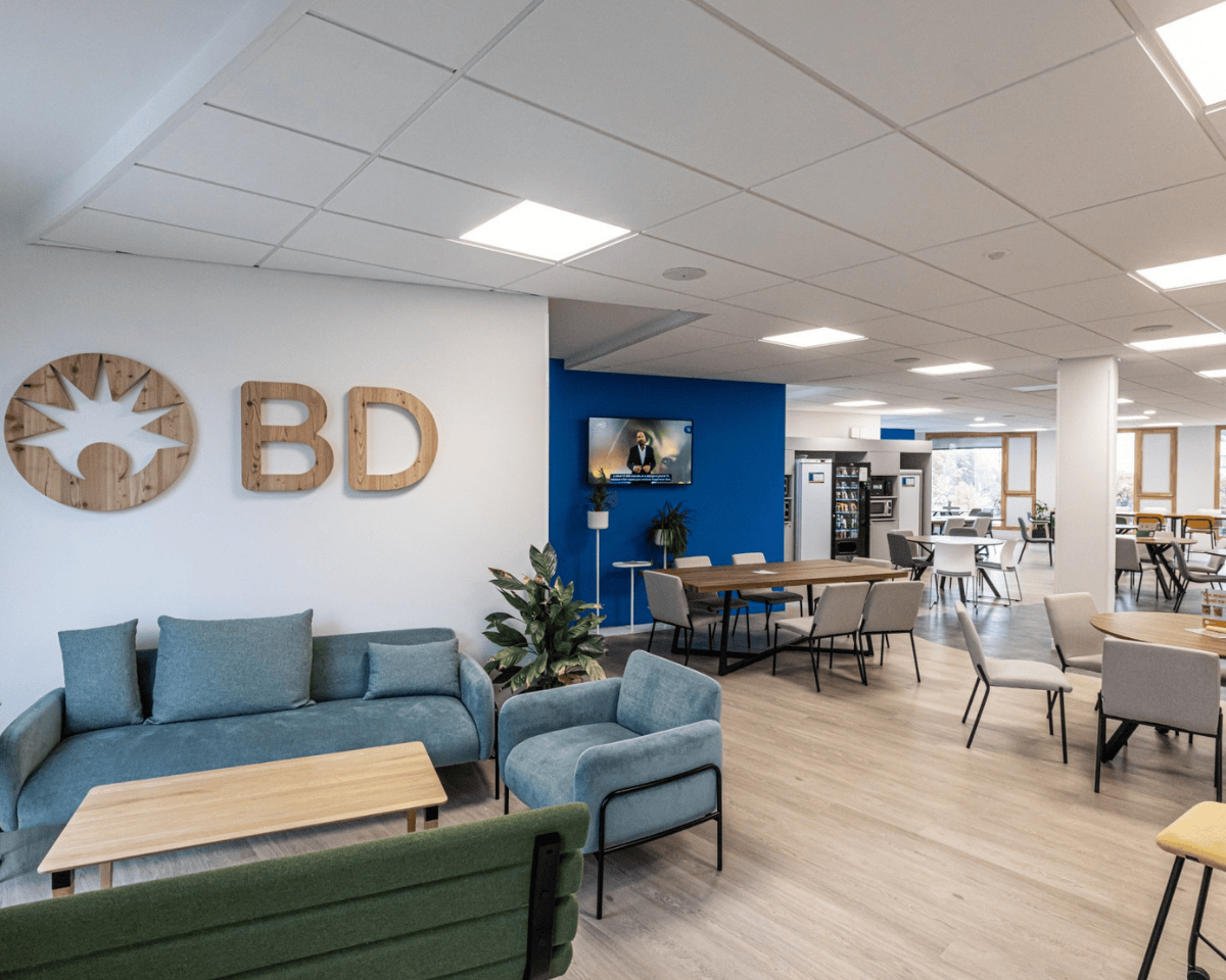 Interior view of a modern BD office lounge featuring seating areas with sofas and chairs, wooden tables, a wall displaying a wooden BD logo, indoor plants, a blue accent wall, and a mounted screen in a bright open workspace.