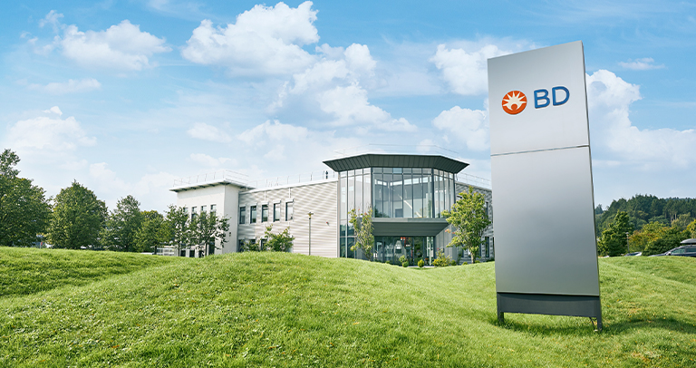 Außenansicht einer BD‑Einrichtung mit einem großen BD‑Schschild im Vordergrund und einem modernen Gebäude mit großen Glasflächen sowie umliegenden Grünflächen unter einem teilweise bewölkten Himmel.