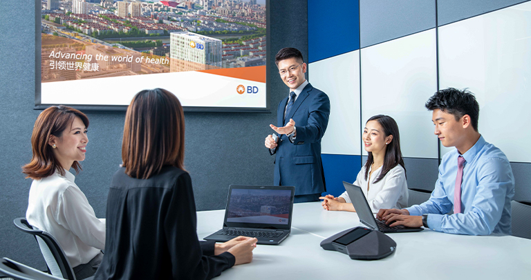 Five individuals seated and standing around a conference table in a modern meeting room, using laptops and looking toward a wall‑mounted presentation screen. The screen displays an image of a cityscape with a BD building.
