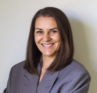 Portrait style photo of an individual with shoulder length dark hair smiling at the camera wearing a grey blazer.