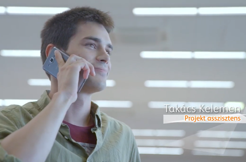 Individual holding a smartphone to the ear inside a bright indoor space with overhead lighting. On-screen text in the lower right corner reads ‘Takács Kelemen’ and ‘Projekt asszisztens.