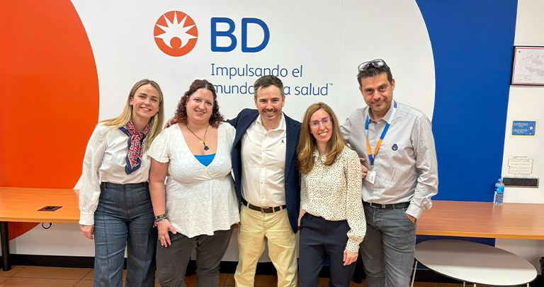 Cinco personas de pie una al lado de la otra frente a una pared con la marca BD que muestra el logotipo de BD y la frase “Impulsando el mundo de la salud”, con mesas de madera colocadas a lo largo de la pared.