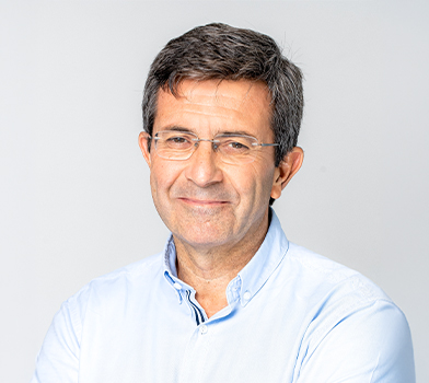 Persona con lentes y una camisa clara con cuello, de pie en un espacio interior frente a un fondo liso de color gris claro, en una fotograf&iacute;a de retrato en primer plano.