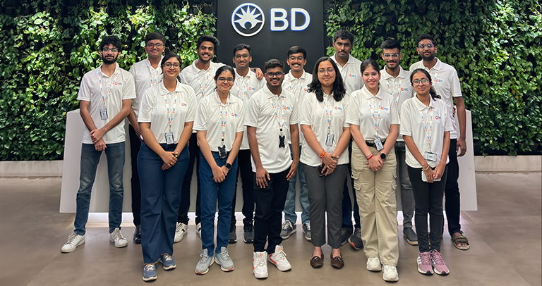 A large group of people smiling directly at the camera in front of a wall with the BD logo that also has foilage growing off it. They are posing in an indoor space.