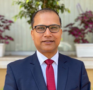 A portrait style image of a subject smiling directly at the camera wearing a navy suit, red tie, white collared shirt and glasses.