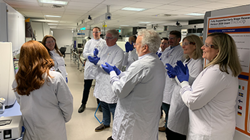 Group of BD associates wearing lab coats and gloves gathered in a laboratory workspace during a team demonstration.