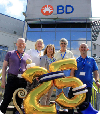 Group of BD associates standing outside a BD building holding a &ldquo;25&rdquo; anniversary display.