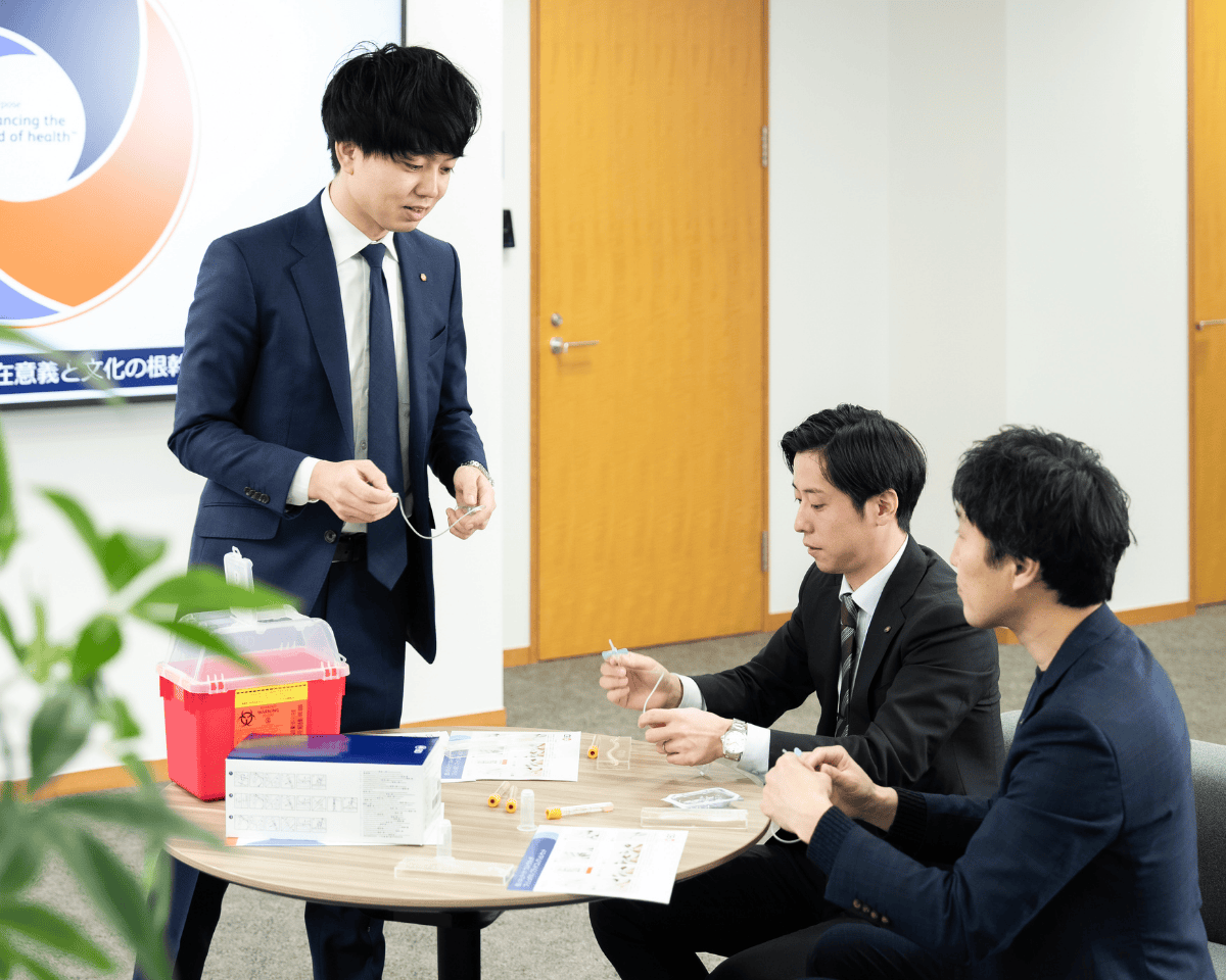 「小さな円形テーブルを囲み、医療トレーニング用のチューブや印刷資料などの教材が置かれたオフィス室内にいる3人の人物。背後には円形グラフィックを表示したスクリーンが見える。」