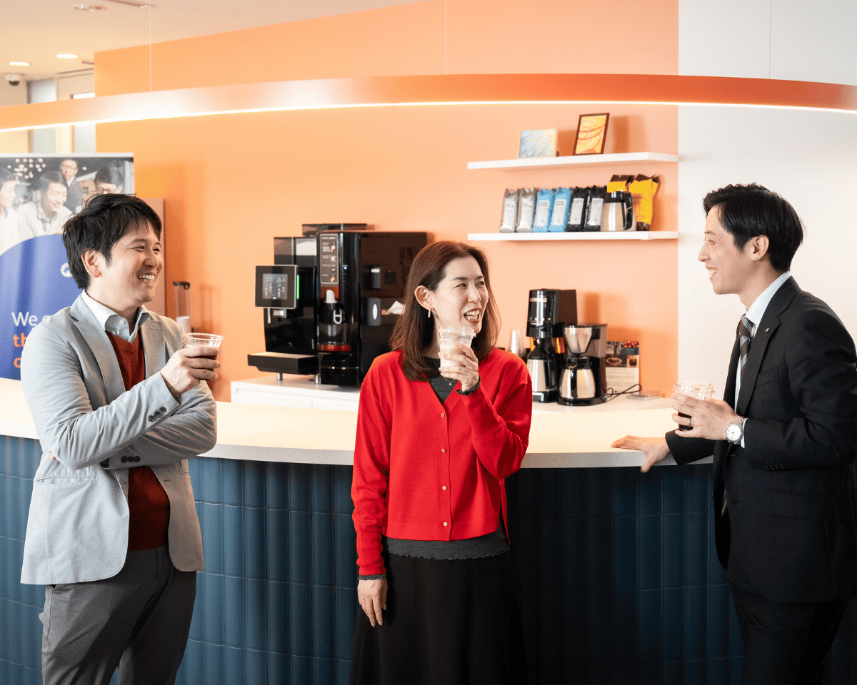 Three individuals standing at an office café counter with beverages in hand. Behind them are coffee machines, shelves with supplies, and an orange accent wall.