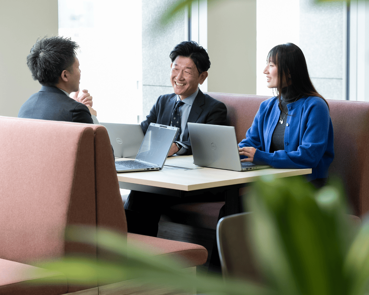「モダンなオフィス空間で、ボックス席のテーブルに着席し、それぞれがノートパソコンを使用している3人の人物。大きな窓から自然光が入り、テーブル周囲には柔らかいシートが配置され、手前には緑の植物が見える。」