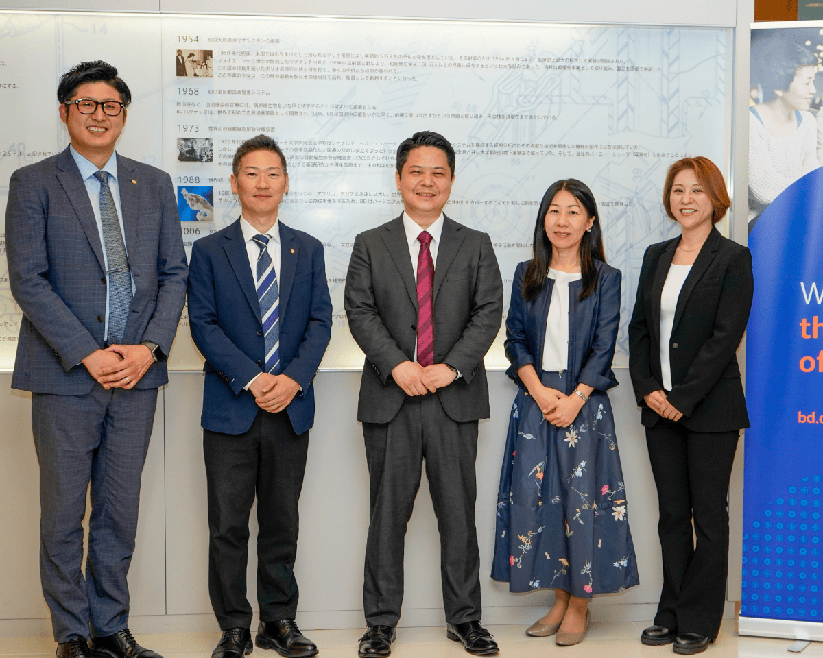 「歴史的なタイムラインの文章と画像が印刷された壁の前に立つ5人の人物。横にはBDブランドの青い縦型バナーが見える。」