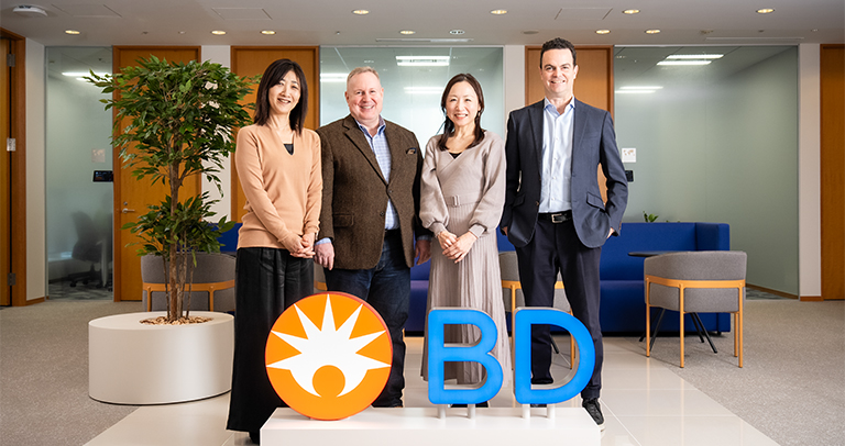 「オフィスロビーで、オレンジ色のBDシンボルと青いBD文字のディスプレイの後ろに立つ4人の人物。背景には、座席エリア、室内植物、明るい色の壁と木材アクセントが見える。」