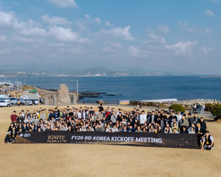 바다와 멀리 보이는 해안선을 배경으로, ‘FY26 BD Korea Kickoff Meeting’이라 적힌 현수막을 들고 해안가 야외에 모여 있는 대규모 동료 그룹.