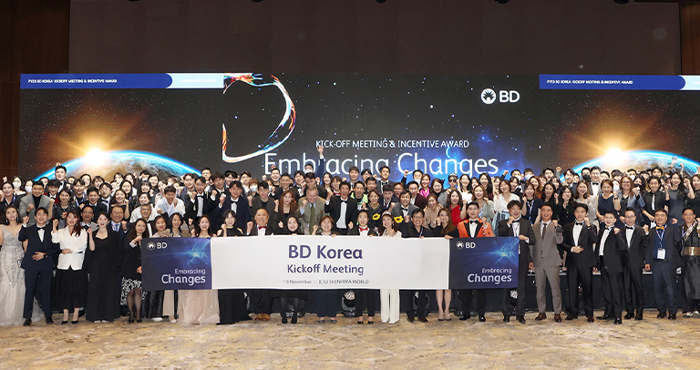“무도회장 분위기에서 정장을 입은 많은 사람들이 모여 있으며, ‘BD Korea Kick Off Meeting’이라고 적힌 표지판을 들고 있다.”