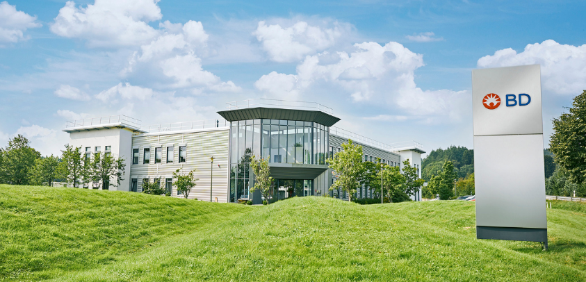 Modern office campus with glass-fronted entrance, white buildings, green lawn, trees, and a BD sign beneath a bright sky.