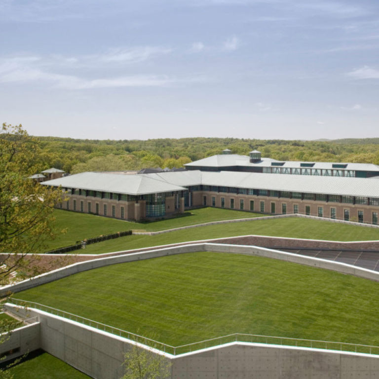 Vista aérea de um complexo de edifícios modernos e de baixa altura, com telhados de cores claras e paredes de tijolos, cercado por extensos gramados verdes e colinas arborizadas e onduladas, sob um céu azul e límpido.