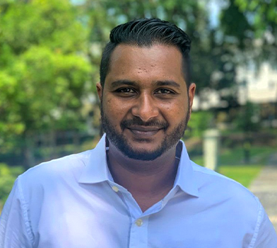 ndividual wearing a white collared shirt standing outdoors in a green, tree‑filled environment with soft natural light and a walkway visible in the background.