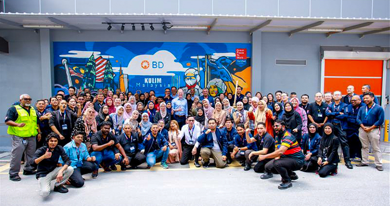A large group of individuals posing together outside a building in front of the BD Kulim Malaysia mural, arranged across the full width of the outdoor area.