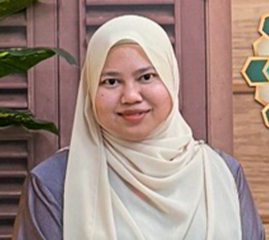 Individual wearing a light-colored headscarf and modest clothing standing indoors in front of wooden paneling, with decorative geometric wall art and a plant in the background.
